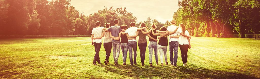 Eine Gruppe von zehn Personen steht mit dem Rücken zur Kamera auf einer Wiese und halten sich.