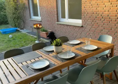 Ein Holztisch mit sechs Stühlen im Außenbereich, gedeckt mit Tellern und einer Pflanze in der Mitte.