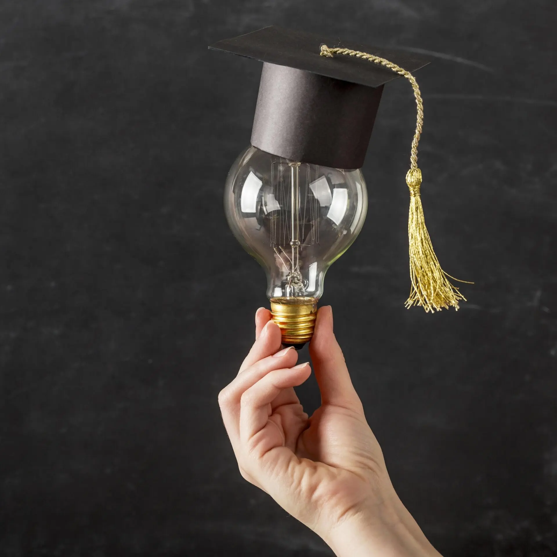 Eine Glühbirne, die von einer Hand gehalten wird, trägt einen schwarzen Absolventenhut mit Quaste.