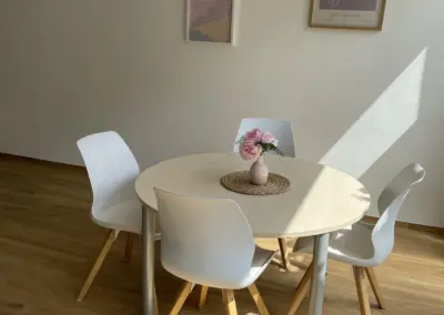 Runder heller Holztisch mit weißen Stühlen, Vase mit rosa Blüten, Sonnenlicht auf Holzfußboden, gerahmte Kunst an der Wand – Kreis Warendorf