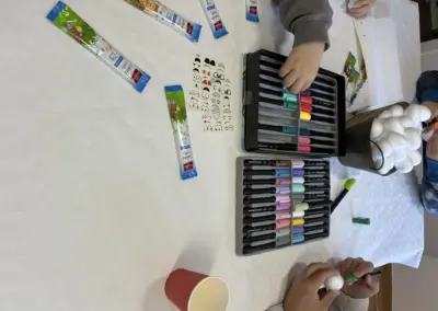 Bunte Wachsmalstifte und Buntstifte in einer Box, Hände bemalen ein Ei; Osterdeko mit grünen und roten Farbtönen, Pastellfarben