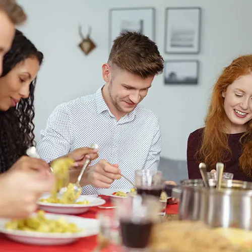 Drei junge Erwachsene essen gemeinsam an einem gedeckten Tisch und greifen mit Besteck zu Pasta und Beilagen.
